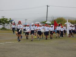 紅白帽と体操服を着用した複数の小学生たちが、白い旗と赤い旗を持って、曇り空の下のグラウンドを行進している様子の写真