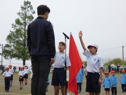 体育服と赤白帽を着用した二人の小学生が、マイクと赤い旗の前で力強く宣誓をしている写真