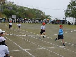 体操服と赤白帽をかぶった小学生たちが、グラウンドの徒競走でゴールテープを切る瞬間を捉えた、運動会の様子の写真