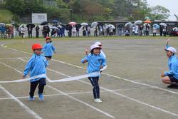 体操服を着た二人の子供が、雨が降るグラウンドでゴールテープを勢いよく切る瞬間を捉えた、運動会の様子の写真