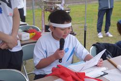 白いTシャツに鉢巻きを巻いた小学生の男の子が、マイクを持って原稿を読み上げている写真