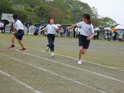赤色の鉢巻きをした二人の小学生が、雨が降るグラウンドでバトンを渡そうとしている、運動会のリレー競技の様子の写真