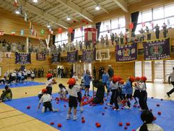 体育館で赤と白の帽子をかぶった子供たちが、青いシートの上で赤玉を集めて玉入れをし、観客席には多くの人が競技を見守っている様子の写真