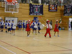 赤い帽子とユニフォームを着た鼓笛隊の子供たちが行進しており、青いユニフォームを着た別のグループの子供たちが楽器を叩いている様子の写真