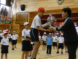 体育館で行われた表彰式で、体育服と紅白帽を着用した生徒が、トロフィーを受け取っている様子の写真