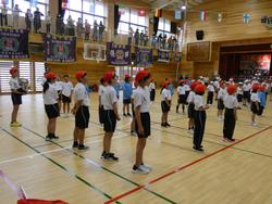 体育館で体操服を着用した複数の小学生たちが、赤い帽子をかぶって整列している写真