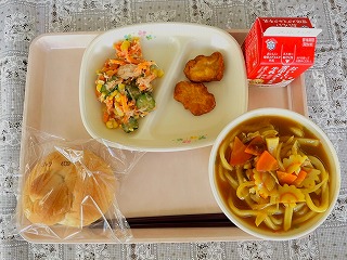 ピンクのトレーに給食が左から時計回りに人参とツナのごまドレッシング、チキンナゲット、牛乳、カレーうどん、バターロールが並んでいる写真