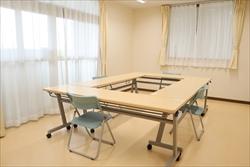 明るいベージュの壁と床の部屋の中央に、可動式の会議机をロの字型に並べ、数脚の椅子が置かれた会議室の写真