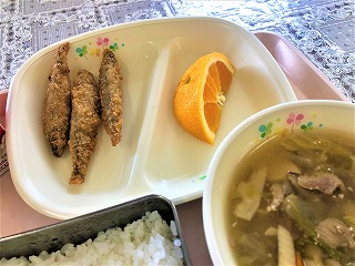 ピンクのトレーに給食が左から時計回りにきびなごフライ（3本）、オレンジ、豚肉と豆腐の中華煮、ごはんが並んでいる写真