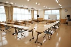 広々とした部屋に、可動式の会議机がロの字型に並べられ、椅子がいくつか置かれた公民館の会議室の写真