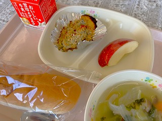 ピンクのトレーに給食が左から時計回りに牛乳、たらのカラフルパン粉焼き、入谷のりんご、きのことケールのポトフ、米粉パンと並んでいる写真