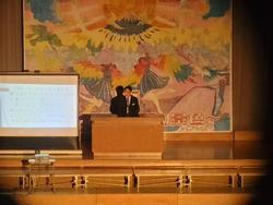 大きな絵が飾られた舞台上の演台に立った男子生徒がマイクを持って発表をし、横のスクリーンに資料が映し出されている様子の写真