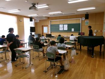 ピアノの置かれた教室で、複数の生徒が机に座って問題を解いており、教室の前方には赤い数字のタイマーが置かれ、先生方が見守る中、算数チャレンジに臨んでいる様子の写真