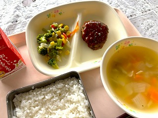 ピンクのトレーに給食が左から時計回りに牛乳、ブロッコリーのソテー、手作りハンバーグ、コンソメスープ、ごはんと並んでいる写真