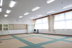 カーペットに幾何学模様がデザインされた広々とした部屋で、窓にはカーテンがかかり、部屋の奥に黒板が設置された公民館の伝統芸能練習室の写真