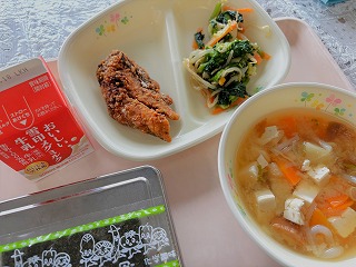 ピンクのトレーに給食が左から時計回りに牛乳、さばのガーリック立田、小松菜と菜花のごまあえ、油ふのみそ汁、味つけのり、ごはんが並んでいる写真