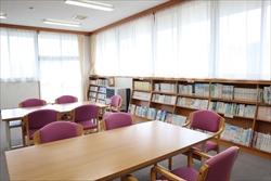 複数の木製テーブルと紫色の椅子が配置され、壁一面の本棚に本がぎっしり詰まった、明るい光が差し込む公民館の図書室の写真