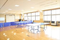 白い壁とフローリングの広い部屋に、青い卓球台が2つ置かれ、壁に大型スクリーンが設置された多目的ホールの写真