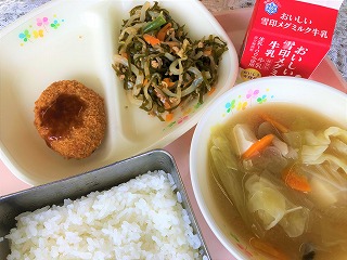 ピンクのトレーに給食が左から時計回りにいかメンチ（パックソース）、すき昆布ともやしの煮物、牛乳、キャベツのみそ汁、ごはんが並べられている写真