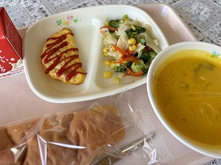 ピンクのトレーに給食が左から時計回りに牛乳、ミートオムレツ、コーンとブロッコリーのソテー、かぼちゃのポタージュ、米粉パンと並んでいる写真
