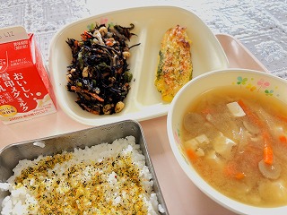 ピンクのトレーに給食が左から時計回りに牛乳、ひじきと大豆の煮物、笹かまの和風ピカタ、ひきな汁、ふりかけごはんが並んでいる写真