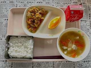 ピンクのトレーに給食が左から時計回りに油ふ丼の具、オレンジ、牛乳、いものこ汁、ごはんと並んでいる写真