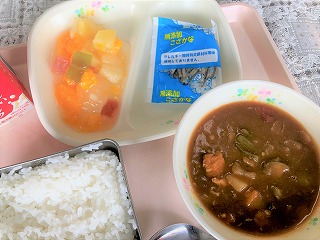 ピンクのトレーに給食が左から時計回りに牛乳、フルーツのゼリーあえ、味付け小魚、夏野菜カレー、ごはんと並んでいる写真