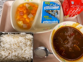 ピンクのトレーに給食が左から時計回りにフルーツのゼリーあえ、味付け小魚、牛乳、夏野菜カレー、麦ごはんと並んでいる写真