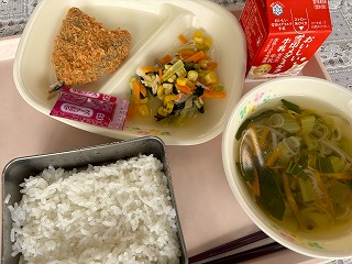 ピンクのトレーに給食が左から時計回りにアジフライ、パックソース、コーンと小松菜のガーリックソテー、牛乳、チンゲン菜のスープ、ごはんと並んでいる写真