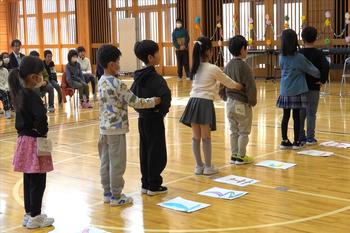 体育館内で低学年の児童が列を作り、前の子の両脇に手を置いて並び、ゲームを行っている写真