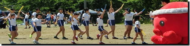 校庭で体操服を着た児童がバトンのようなものを両手に持って踊っており、右側には鉢巻を巻いた赤いオクトパス君のぬいぐるみがいて、奥には木々の緑とテントが並ぶ運動会の賑やかな様子の写真