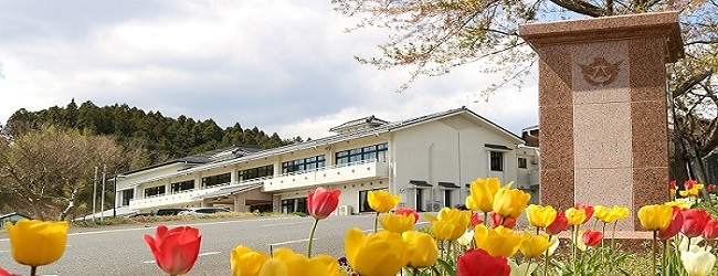 赤や黄色のチューリップが咲く花壇のそばの校門から白い校舎を望む春の学校の風景の写真