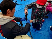 野外のブルーシートの上で赤白帽を被った児童が青の手袋をした方と一緒に制作をしている様子の野外活動の写真