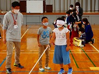 体育館でアイマスクをした小学生が、白い杖を持って歩いており、教員と他の児童がそれを見守っている福祉体験授業の様子の写真