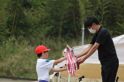 赤い帽子をかぶった男子児童が、演台に立つの関係者の男性からトロフィーを受け取っている写真