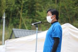 青いTシャツに黒い長袖を重ね着し、白いマスクを着けた関係者の男性がスピーチをしている様子を横から写した写真