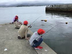 赤い帽子をかぶった子どもたちが海辺の堤防に座り、釣り竿を持って魚釣りをしている写真