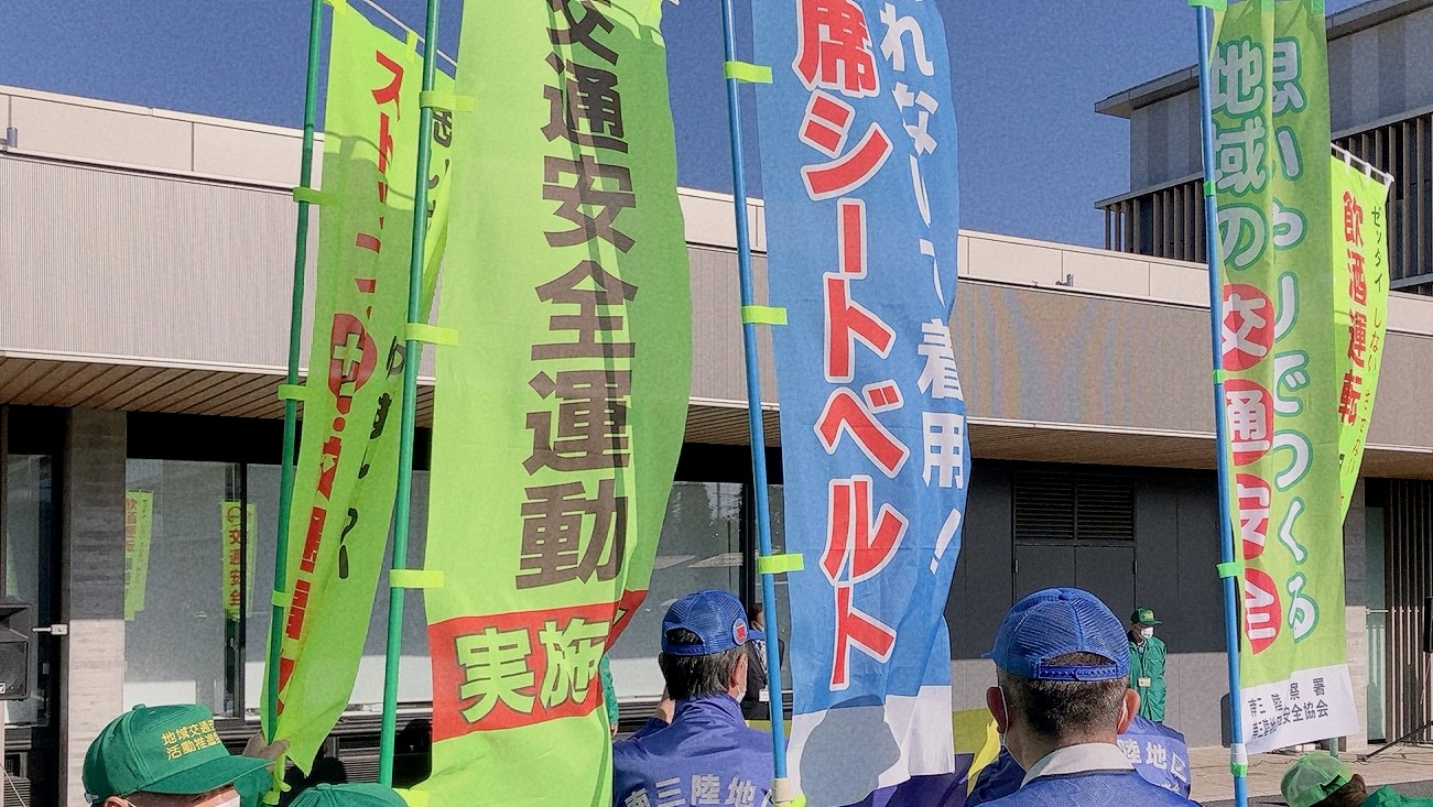 緑や青の帽子をかぶった関係者達が交通安全運動の幟を持って並んだ出動式の様子の写真