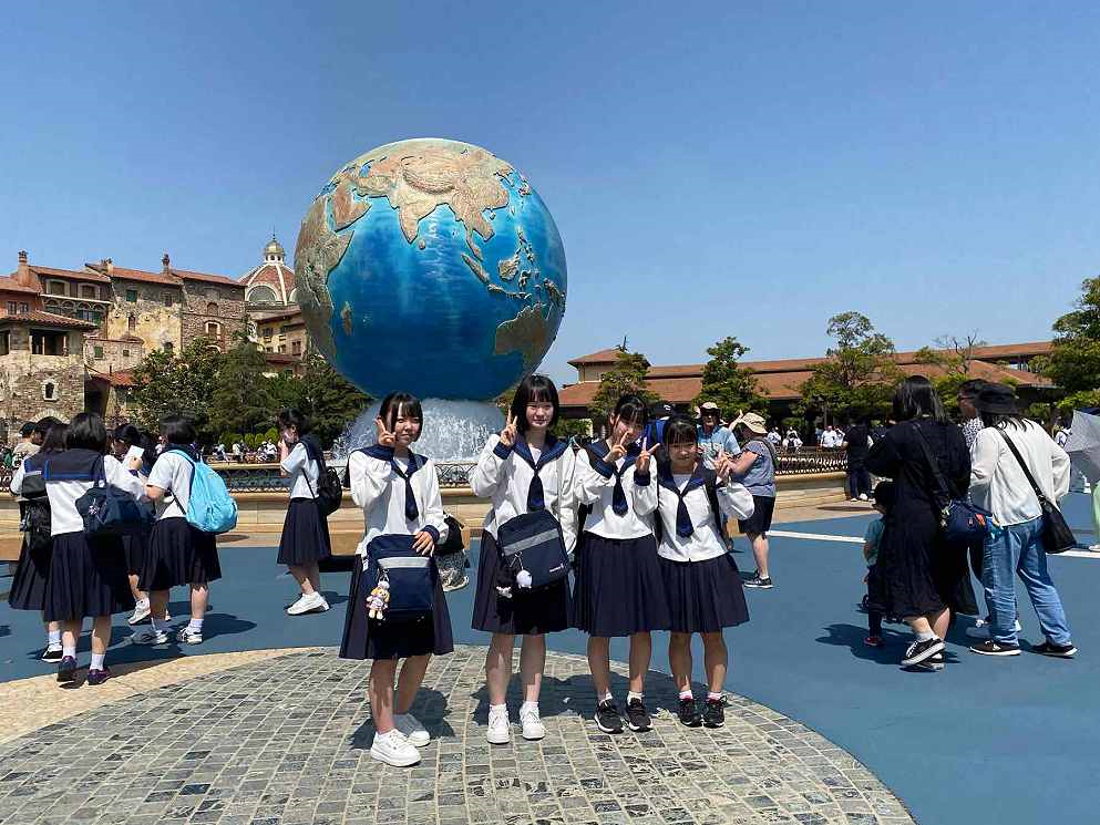 ヨーロッパ風の建物や木々の手前にある、たくさんの人でにぎわう広場の大きな地球儀のオブジェの噴水の前で、制服姿の女子生徒4人が並んでピースサインをしながら記念撮影をしている写真