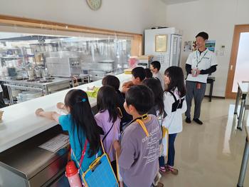 男性の説明を受けながらガラス越しに見える調理場を見学している児童たちの写真