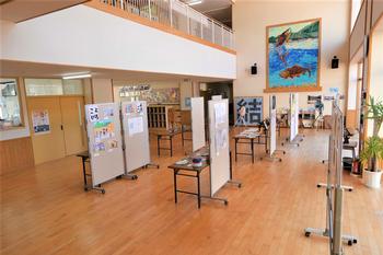 窓から光が差し込む広い廊下に、絵や書道作品が展示されたパネルや机が並んでいる展示品の写真