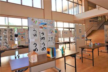 大きな窓から光が差し込む広い廊下で、書道や工作、絵などの児童の作品が机やボードに並べて展示されている写真