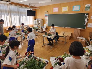 教室に机を半円のように並べ、新聞紙をひいてたくさんの草花を並べフラワーアレンジメントをしている子供たちの写真