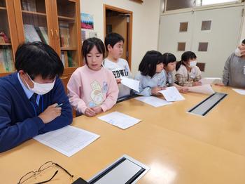 大きな机に座っている先生に子供たちが回答用紙をもって並んでいる室内を写した写真