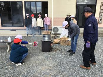 赤白帽を被った子供たちが薪を燃やし、近くで男性が見守りながらその煙が立ち上るかまどで調理をしている屋外炊飯イベントの様子写真