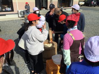 紅白帽をかぶった小学生たちが臼を囲んで、きねで餅つきを楽しむ行事の様子を捉えた写真