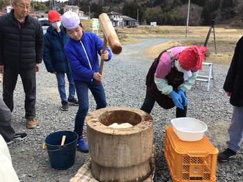 複数の子供と大人が集まり、屋外で大きな臼を使い、男の子がきねを振りかぶって餅つきをしている様子を捉えた写真