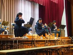 舞台とその前に設置された段の上に並んで座った複数の生徒が、楽器を演奏している写真
