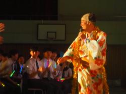 浴衣とちょんまげのカツラを身につけた人物が舞台でパフォーマンスを行い、それを生徒たちがサイリウムを振って見ている写真