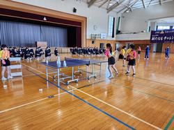 体育館の舞台前に並んだ椅子に制服を着たたくさんの生徒たちが座って見学する中、手前に置かれた卓球台で、ユニフォームを着た女子部員たちが卓球のパフォーマンスを披露している様子の写真